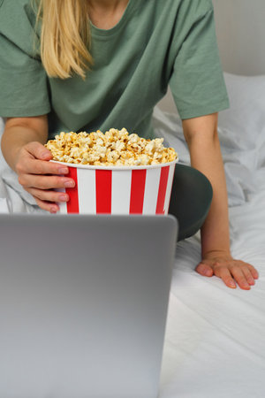 Popcorn bucket in the hands of a young girl preparing to watch a movie in bed on the laptop. Showtime. Eating delicious sweet snacks. Watching new film in homeの写真素材