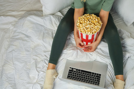 Popcorn bucket in the hands of a young girl preparing to watch a movie in bed on the laptop....
