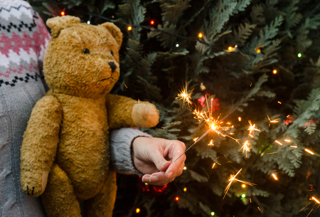 Christmas scene - woman in knitted sweater holding an old teddy bear and sparkler before christmas treeの写真素材