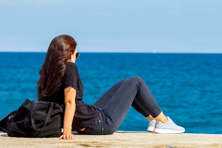 Young girl sitting on the edge of the pier and watching the blue sea.の写真素材