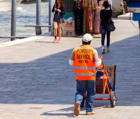 Venice, Italy - 25 May, 2015: Porter with a trolley in a bright orange vest. A hotel employee on the transport of luggage on the Venetian quay.のeditorial素材