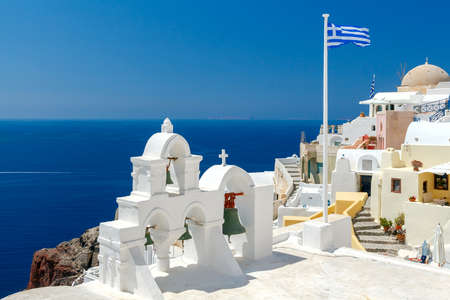 White tower with bells and the Greek flag on a background of blue sea and sky in the village of Oia. Santorini, Greece.の写真素材