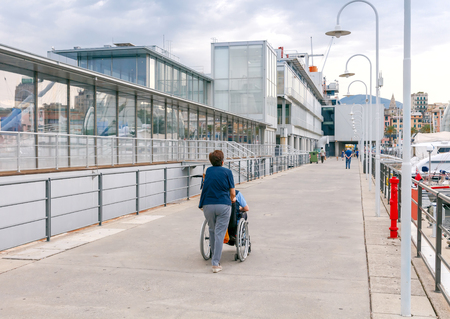 A woman carries a disabled person in a wheelchair on the waterfront in the city of Genoa.の写真素材