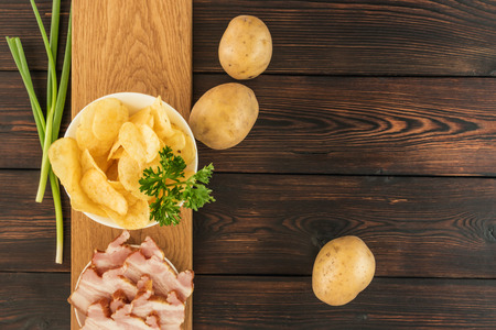 Still life with chips and raw potatoes, bacon and green onions on a dark wooden backgroundの写真素材