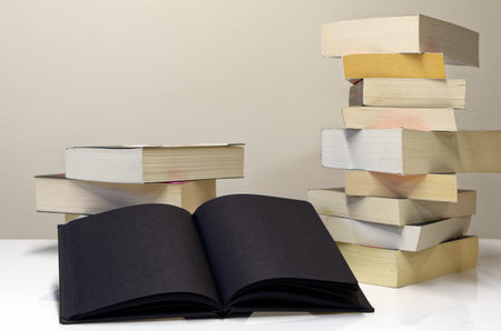 Black open book in foreground of two piles of books on the white table and light grey background