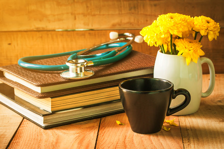 selective focus of empty cup of coffee on wooden floor with yellow flower in white pot and stethoscope on books , morning sunlight. vintage color tone.の写真素材