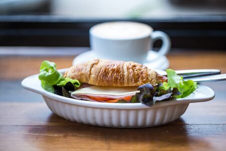blurred of Ham Cheese Croissant with fresh vegetable in white bowl and hot latte art coffee on wooden table in coffee shop.の写真素材