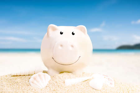 summer happy piggy bank on sand beach over blurred tropical blue sea background, image saving vacation concept.の写真素材