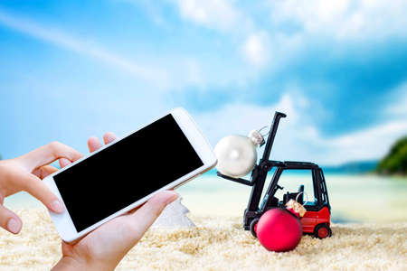 woman hand hold and touch screen smartphone or cellphone over Christmas decorations on white sand beach.の写真素材
