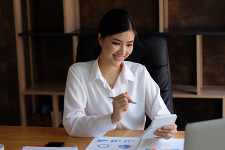 Beautiful asian businesswoman using smart phone and working with financial documents at office.の写真素材