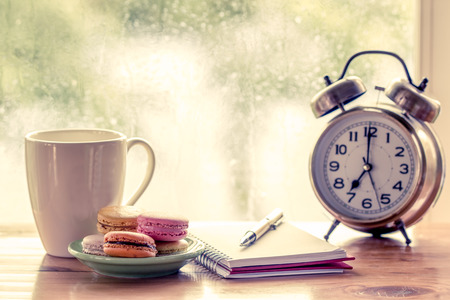 Colorful macaroons and coffee cup with pen on notebook and alarm clock on rainy day window background in vintage color toneの写真素材