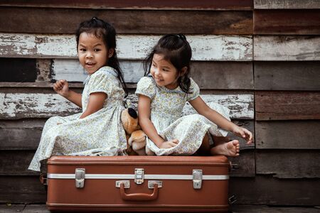 children two cute asian little girls are sitting on suitcase and playing together on wooden background in vintage retro styleの写真素材