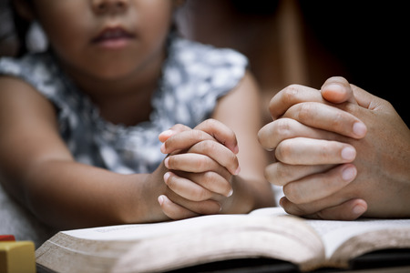 Mother and little girl hands folded in prayer on a Holy Bible together  for faith concept in vintage color toneの写真素材