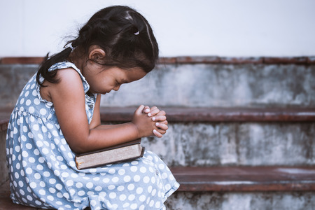Cute asian little girl closed her eyes and  folded her hand in prayer on a Holy Bible for faith concept in vintage color toneの写真素材