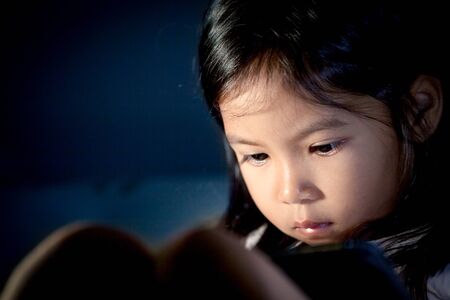 Cute asian little girl using digital tablet at night in the bedroom in dark blue color toneの写真素材
