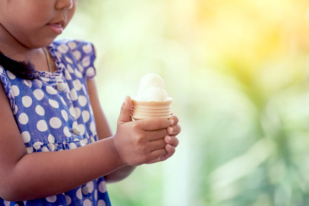 Cute asian little girl is holding melting ice cream waffle cone in hand in green nature background