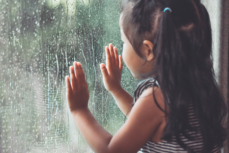 Sad asian little girl looking outside through the window in the rainy day in vintage color toneの写真素材