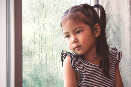 Sad asian little girl looking outside through the window in the rainy day in vintage color toneの写真素材