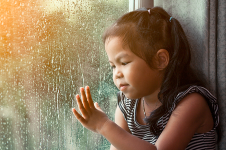 Sad asian little girl looking outside through the window in the rainy day in vintage color toneの写真素材