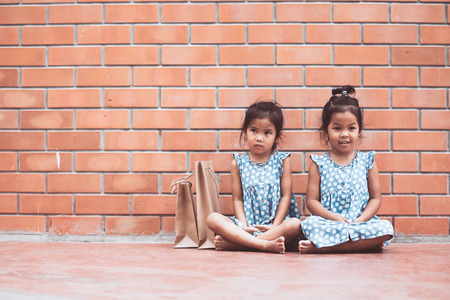 Two asian kid girls sitting on the floor and waiting their parent shopping on brick wall background in vintage color toneの写真素材