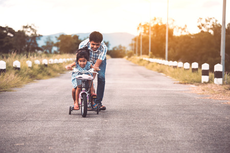 Asian kid girl having fun to ride bicycle with father help to pushing them together in the park in vintage color toneの写真素材