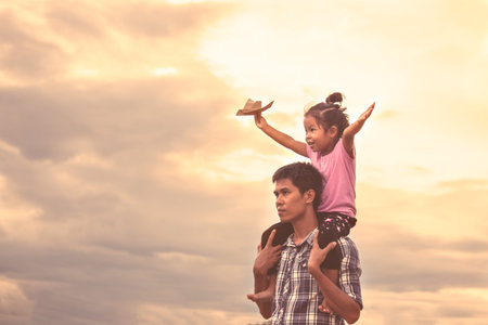 Cute asian girl riding on father's shoulder and playing with toy paper airplane against sky in vintage color toneの写真素材