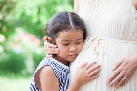 Asian child girl  listening baby and hugging pregnant mother's  belly with love waiting for new baby sisterの写真素材
