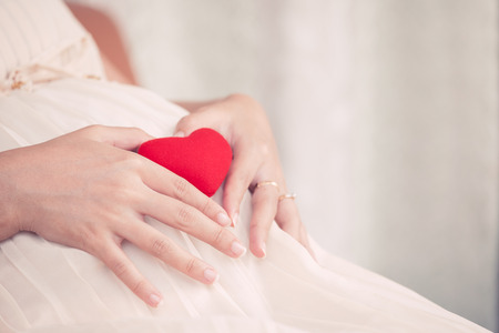 Pregnant woman sitting and making hand heart shape with red heart on her big belly, mother love babyの写真素材