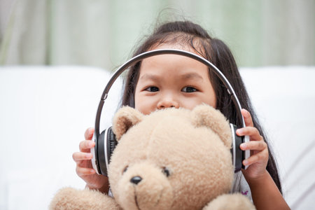 Happy asian child girl playing with her teddy bear with headphones in her roomの写真素材