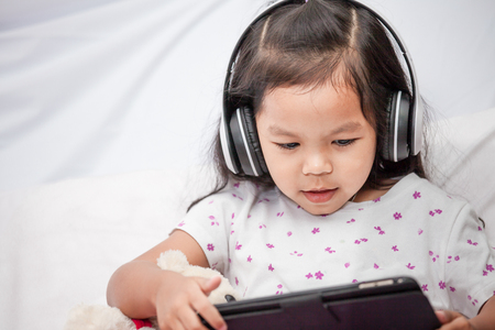 Cute asian little child girl in headphones is using a tablet in her roomの写真素材