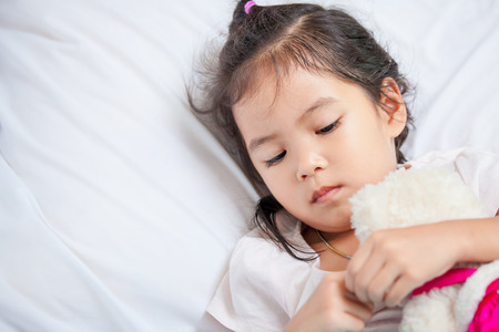 Cute asian little child girl hugging her teddy bear and lying in bed with sad moody at homeの写真素材