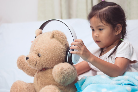 Cute asian child girl playing with her teddy bear with headphones in her roomの写真素材