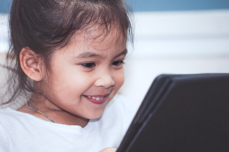 Happy asian child girl sitting on sofa smiling  and  using smart phone, cellphone,tablet in the roomの写真素材