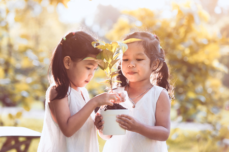 Two asian little child girls holding young tree in pot together for prepare plant on ground as save world concept in vintage color toneの写真素材