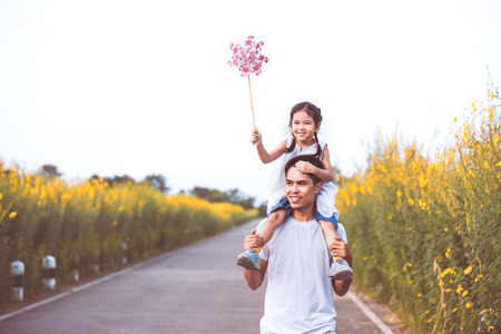 Cute asian little girl playing with wind turbine and riding on father's shoulders in the flower garden in vintage color tone. Father and daughter having fun to play together.の写真素材