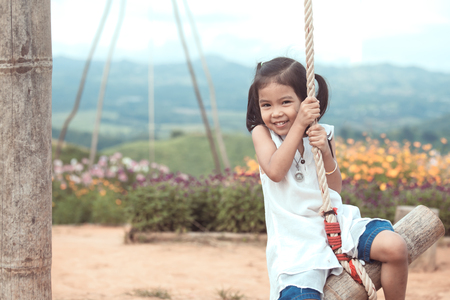 Happy asian little child girl having fun to play on wooden swings in playground at the gardenの写真素材