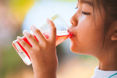 Cute asian little child girl drinking red juice water with ice from glass in the summer timeの写真素材