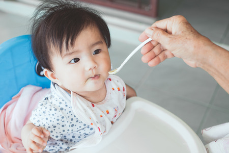 Grandmother feeding cute asian baby girl with a spoon at home. Little baby girl sitting in the chair and eating her meal with innocence.の写真素材