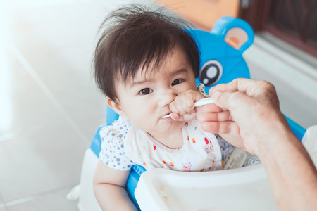 Grandmother feeding cute asian baby girl with a spoon at home. Little baby girl sitting in the chair and eating her meal with innocence.の写真素材