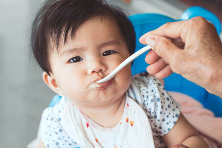 Grandmother feeding cute asian baby girl with a spoon at home. Little baby girl sitting in the chair and eating her meal with innocence.の写真素材