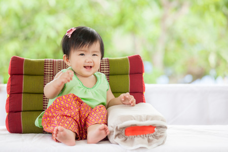 Cute asian baby girl in Thai traditon dress sitting with pillow smiling with happinessの写真素材