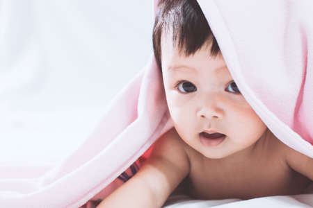 Cute asian baby girl smiling under pink blanket and lying on her stomach on the bedの写真素材