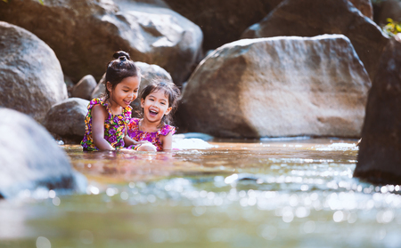 Two asian little child girls sitting on the rock and playing water in the riverside together in summer time with fun and happinessの写真素材