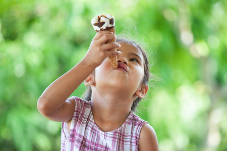 Cute asian little child girl is eating delicious icecream cone with fun and happinessの写真素材