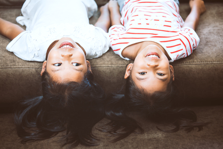 Two cute little child girls are lying on their backs on sofa and playing together at home with fun and happinessの写真素材