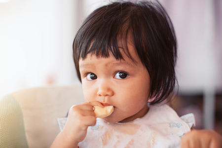 Cute baby asian child girl eating healthy food by herself and making a mess on her face and handの写真素材