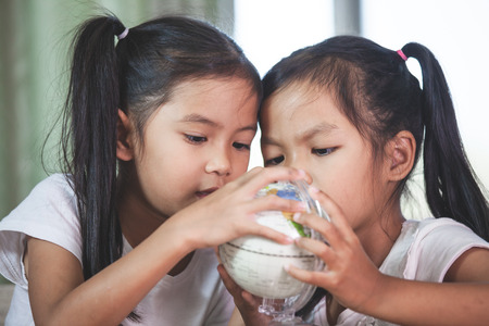 Two cute asian child girls use magnifier to look and study at the globe together in classroomの写真素材