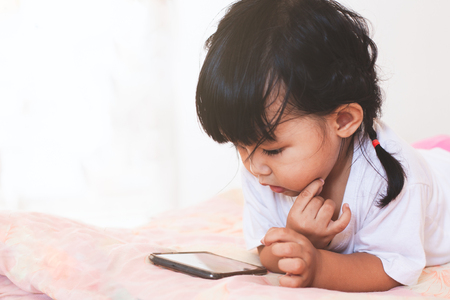 Cute asian baby girl playing smartphone lying on her bed in her roomの写真素材