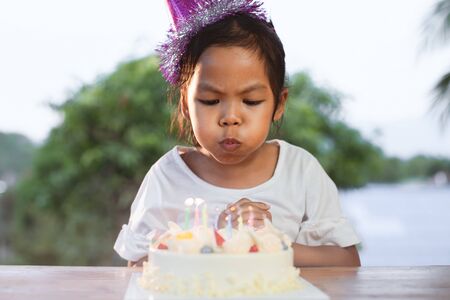 Cute asian child girl celebrating birthday and blowing candles on birthday cake in the partyの写真素材