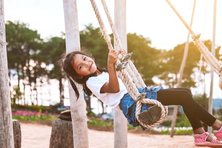 Happy asian child girl having fun to play on wooden swings in playground with beautiful natureの写真素材
